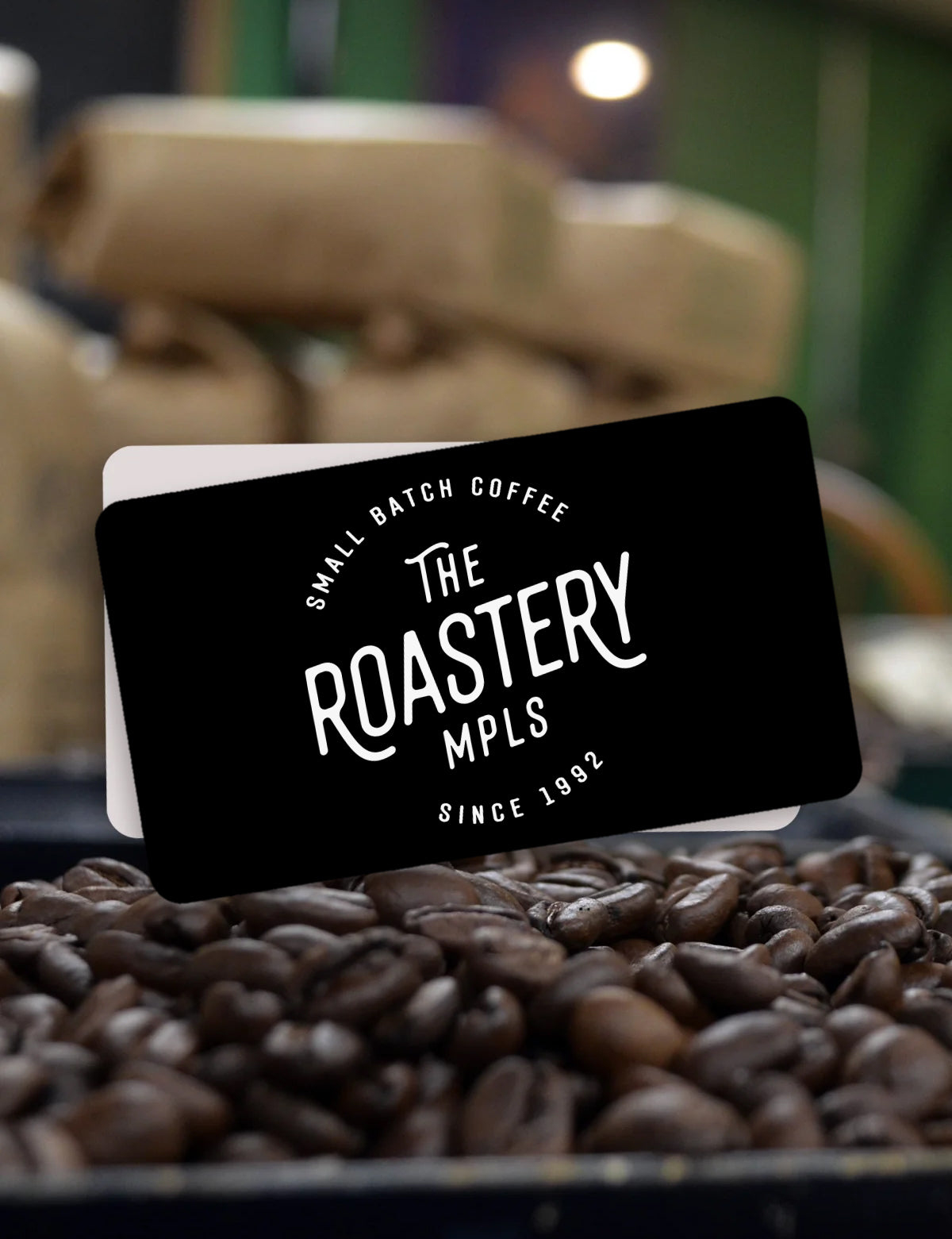 The Roastery Mpls coffee gift card on a bed of coffee beans with blurred coffee bags in the background.